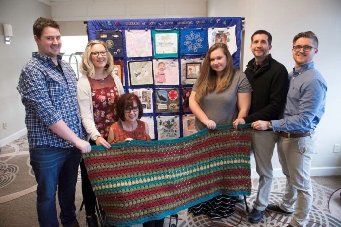 Family members hold a handmade wrap and stand before the "Threads of Love" Memorial Quilt.