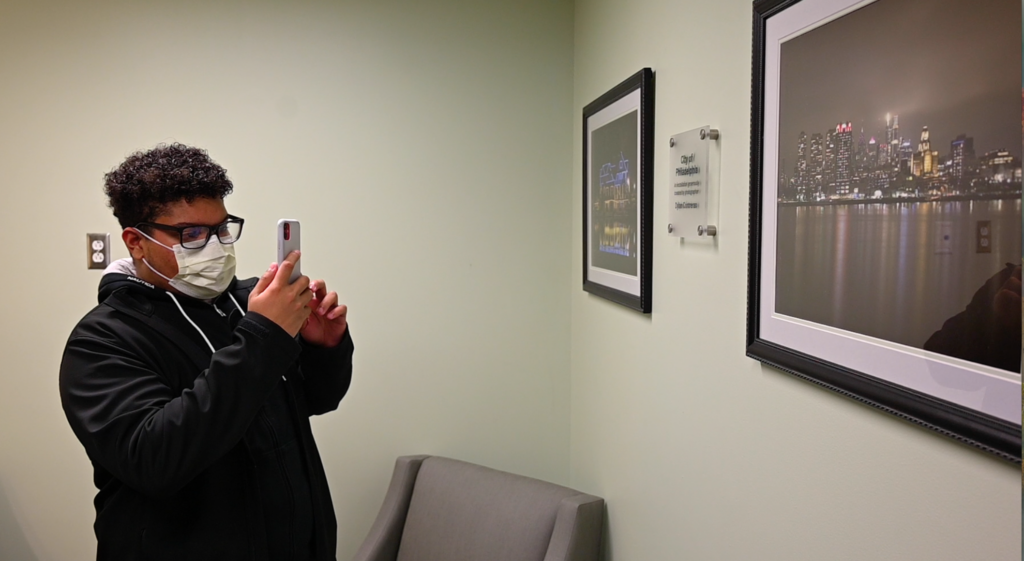 A young man takes photos with his cellphone of artwork hanging on the wall of a room.