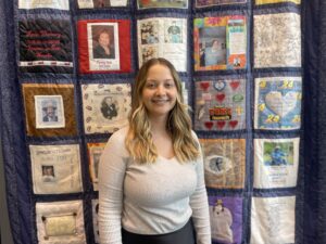A woman stands in front of a quilt to pose for a photo. 