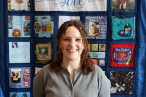 A woman smiles for a photo in front of a quilt honoring donor heroes.