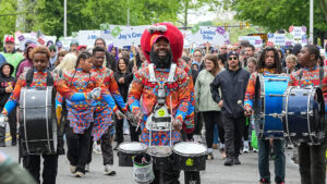 Drummers in bright patterned outfits march down a city street, smiling, with a cheering crowd and signs behind.