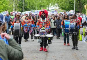 Drummers in bright patterned outfits march down a city street, smiling, with a cheering crowd and signs behind.