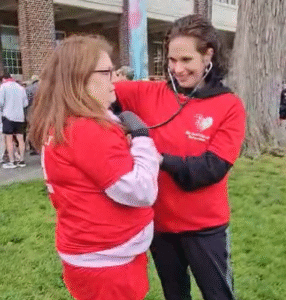 Two women in red volunteer shirts outdoors; one uses a stethoscope to check the other's blood pressure on her arm.