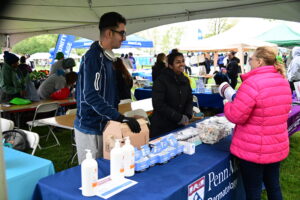 People at a community health fair under tents; a booth hands out skincare products on a table with pumps and boxes in view.