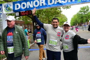 Runners smile and raise their arms at the finish line under a blue DONOR DASH banner; bibs show 483, 1397, 965.