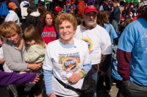 Smiling woman wearing a white 'Mother of the Dash' T‑shirt stands at a crowded finish-line event among spectators and volunteers.