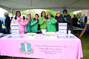 Group of smiling women under a white canopy at a campus event, standing behind a pink table with brochures for Alpha Kappa Alpha Sorority.