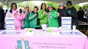 Group of smiling women under a white canopy at a campus event, standing behind a pink table with brochures for Alpha Kappa Alpha Sorority.