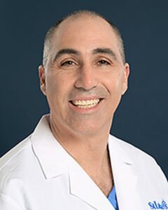 Smiling male doctor in a white coat against a blue background, looking at the camera.