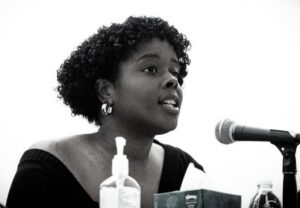 A woman speaks into a microphone at a panel, with a bottle and small items on the table in front of her.