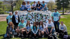 Group of volunteers posing on a painted rock sculpture with a tree mural in a grassy park on a sunny day, many wearing teal Donate Life shirts.