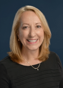 Professional headshot of a smiling blonde woman in a black top against a blue background.