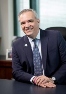 Smiling middle-aged man in a navy suit sits at a desk in an office with a blurred window in the background.