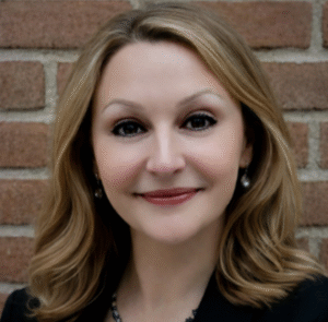 Head-and-shoulders portrait of a smiling woman with light brown hair, wearing a dark blazer, against a brick wall background.