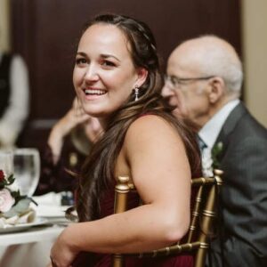 Smiling woman with long dark hair in a burgundy dress seated at a banquet table, with an older man in a suit in the background.