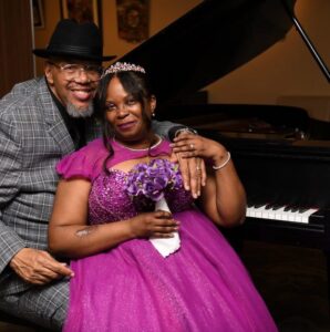 A smiling couple sits close together by a grand piano; the woman in a purple gown holds a bouquet and wears a tiara, while the man in a gray checkered suit and hat rests his arm around her.