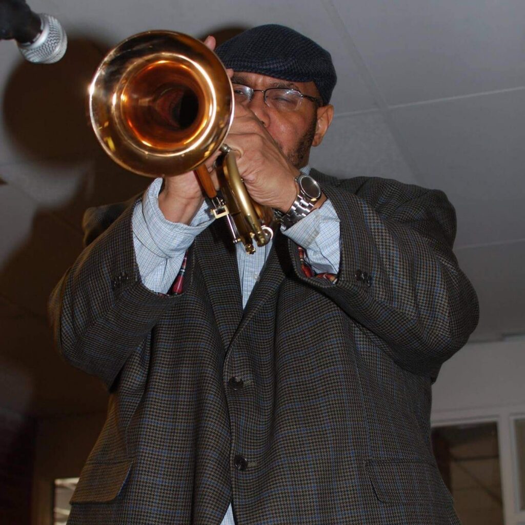 Man wearing a cap and glasses playing a brass trombone up close indoors, microphone nearby.