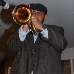 Man wearing a cap and glasses playing a brass trombone up close indoors, microphone nearby.