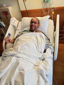 Man in a hospital bed smiling at the camera, covered with white sheets and wearing a hospital gown, with monitors and medical equipment in the background.