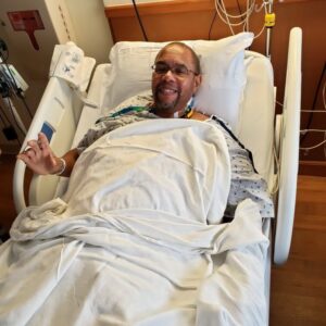 Man in a hospital bed smiling at the camera, covered with white sheets and wearing a hospital gown, with monitors and medical equipment in the background.