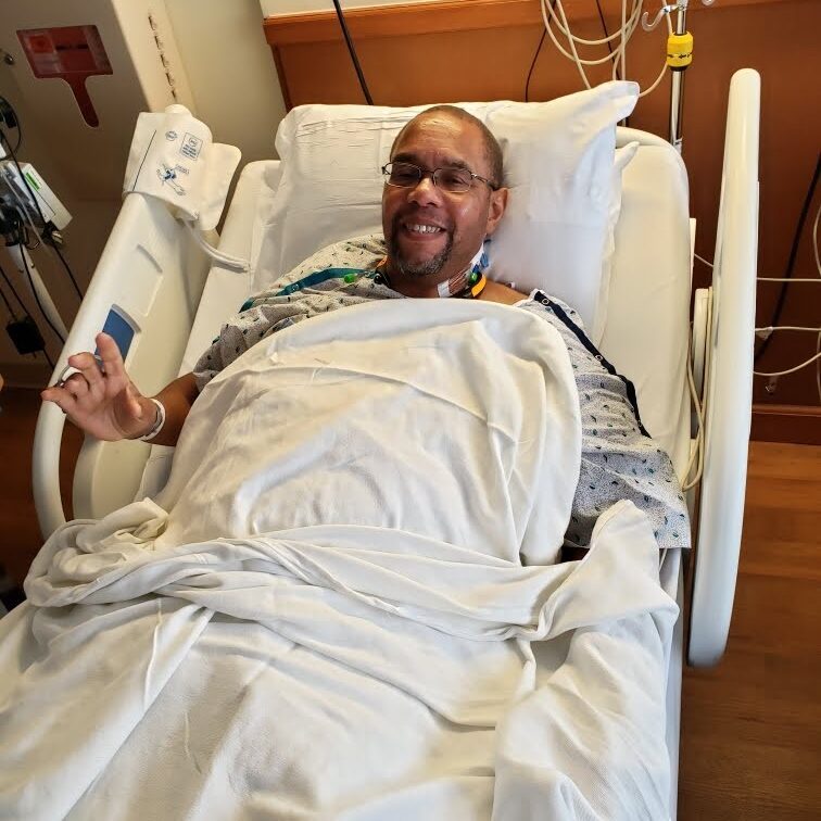 Man in a hospital bed smiling at the camera, covered with white sheets and wearing a hospital gown, with monitors and medical equipment in the background.